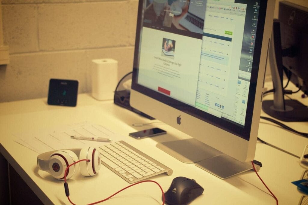 A desktop computer with a large monitor, keyboard, mouse, white Beats headphones, smartphone, and scattered papers on a white desk in an office setting. A website is open on the screen.