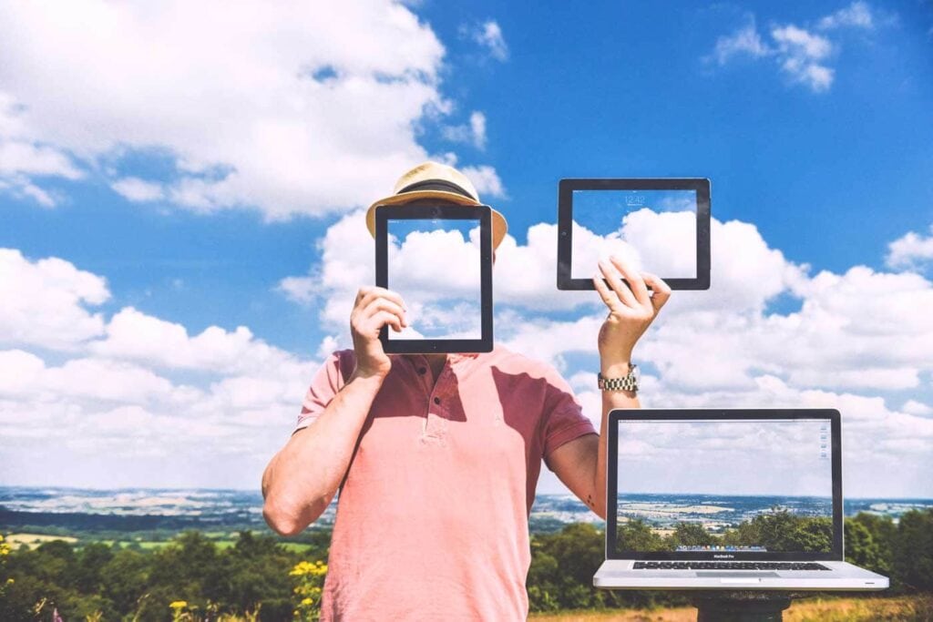 A person holds a tablet in front of their face and another in one hand, with a laptop in front; each screen displays the matching sky and clouds, blending with the real background.