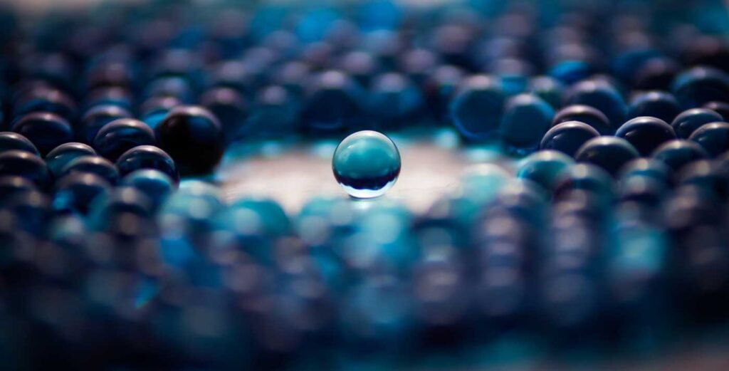 A single clear blue marble is centered and in focus, surrounded by many blurred blue marbles, creating a striking visual contrast between the sharp and soft elements.