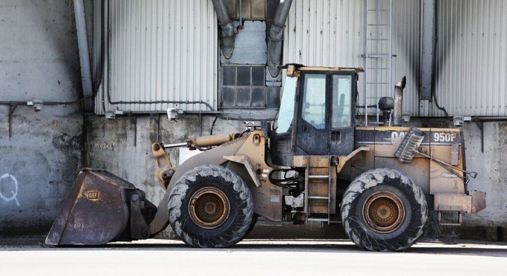 A large, dusty front-end loader with a bucket attachment is parked on concrete in front of a corrugated metal wall and a window.