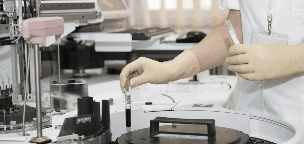 A person wearing gloves holds test tubes in a laboratory, surrounded by scientific equipment and machines.