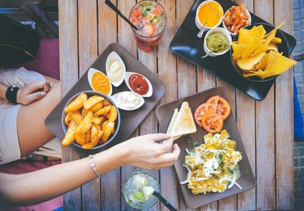 Overhead view of a wooden table with potato wedges, sauces, nachos with dips, a salad, drinks, and a hand reaching for food.