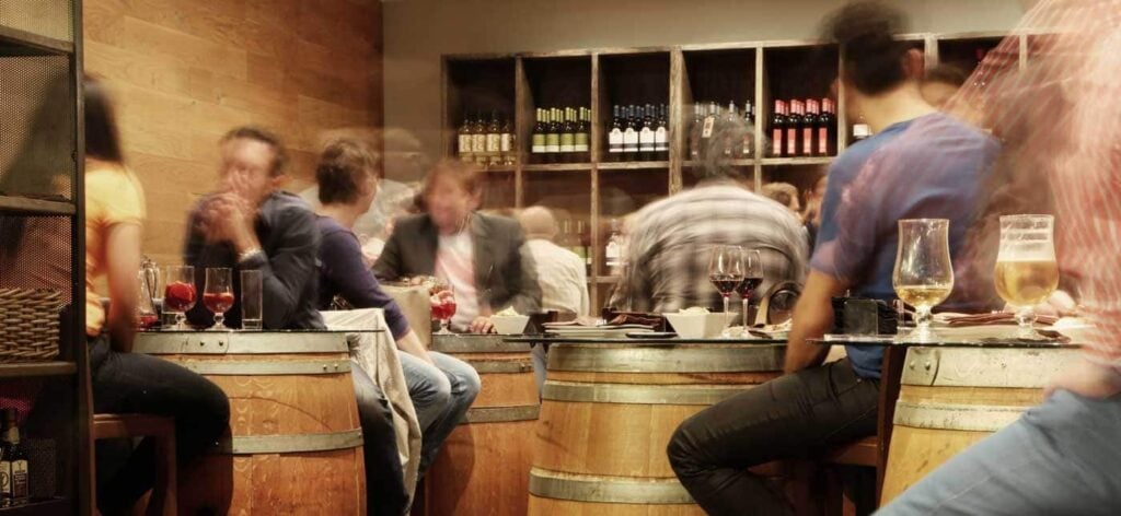 People sit around tables made from barrels in a cozy bar or restaurant, enjoying drinks. Shelves of wine bottles are visible in the background. The atmosphere appears lively and social, with a mix of blurred movement.
