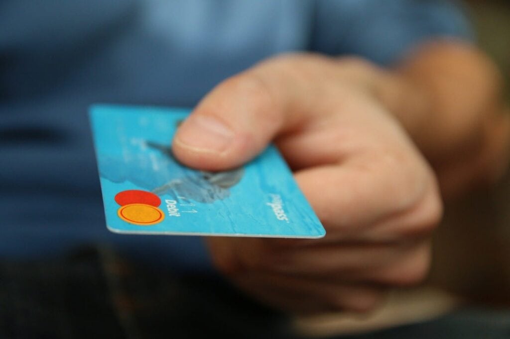 A person holds out a blue debit card, extending it forward with their hand, as if offering it for payment or transaction. The card’s chip and Mastercard logo are visible.