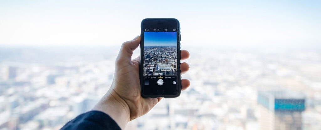 A hand holds a smartphone, taking a photo of a cityscape from a high vantage point, with the blurred city visible in the background.