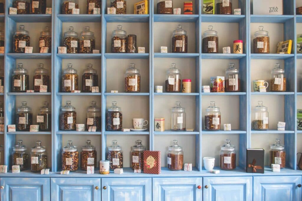 Shelves with blue trim filled with glass jars containing various loose teas, each labeled. Some shelves also hold colorful mugs, boxes, and decorative items. The lower cabinets are closed.