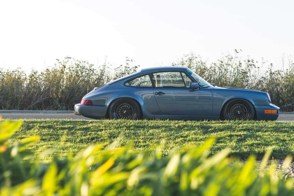 A classic blue coupe car parked on the side of a road, with greenery and tall grass in the background, under a clear sky.