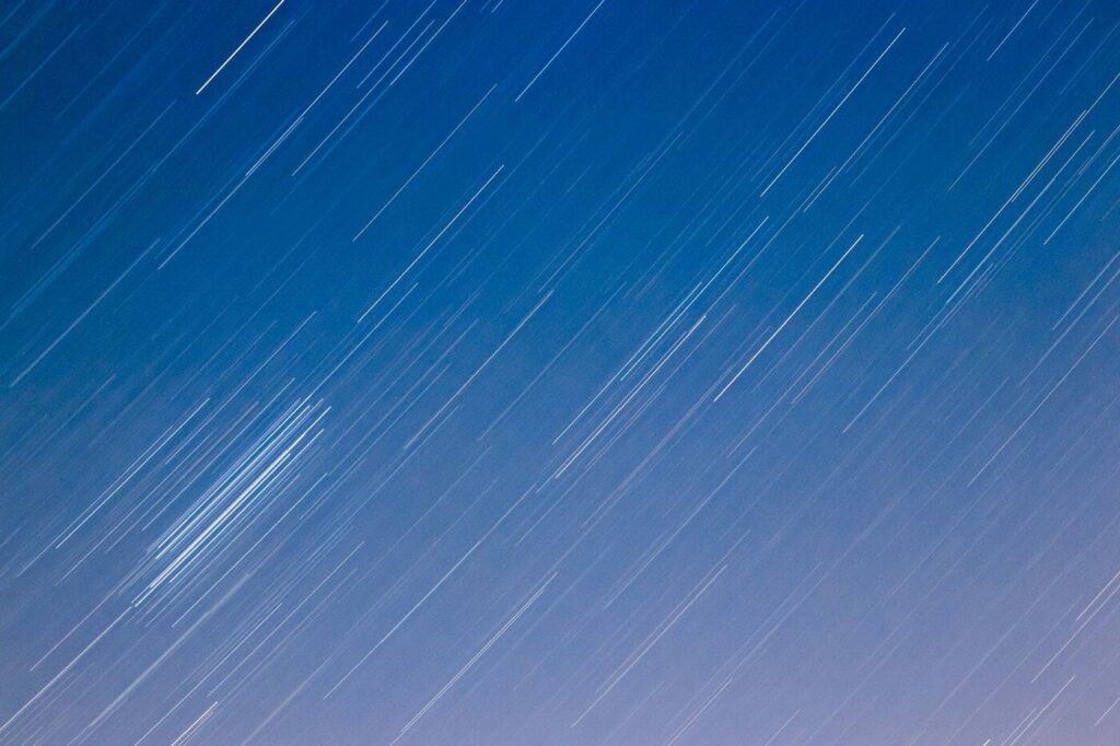 Blue night sky with numerous white streaks showing star trails, created by long-exposure photography. The lines move diagonally across the image, giving a sense of motion and the Earth’s rotation.