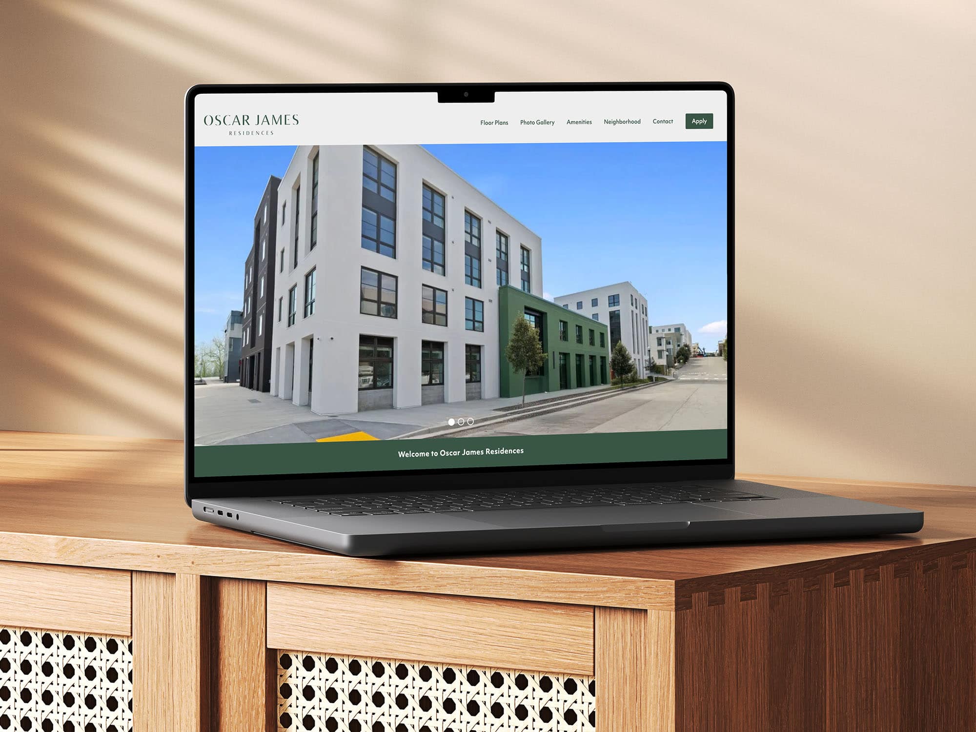 A Laptop On A Wooden Cabinet Displays A Website For Oscar James Residences, Showing A Modern Apartment Building With Large Windows And White And Gray Exterior Walls.
