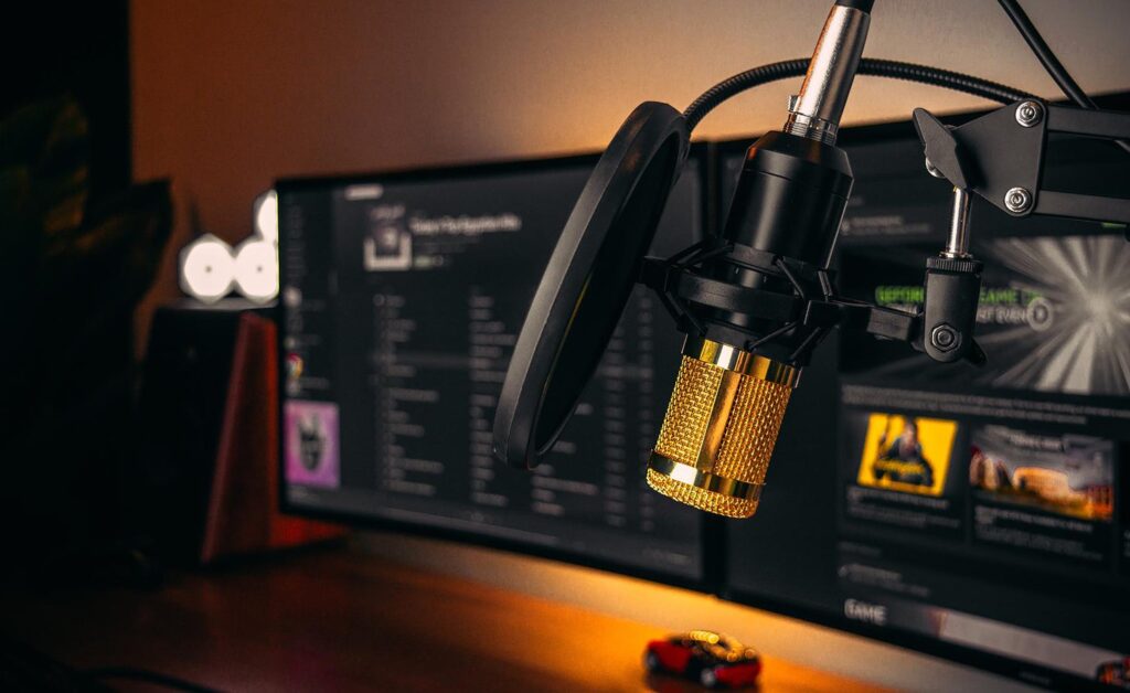 A gold and black microphone with a pop filter is mounted on a desk arm in front of computer monitors displaying various screens in a dimly lit room.