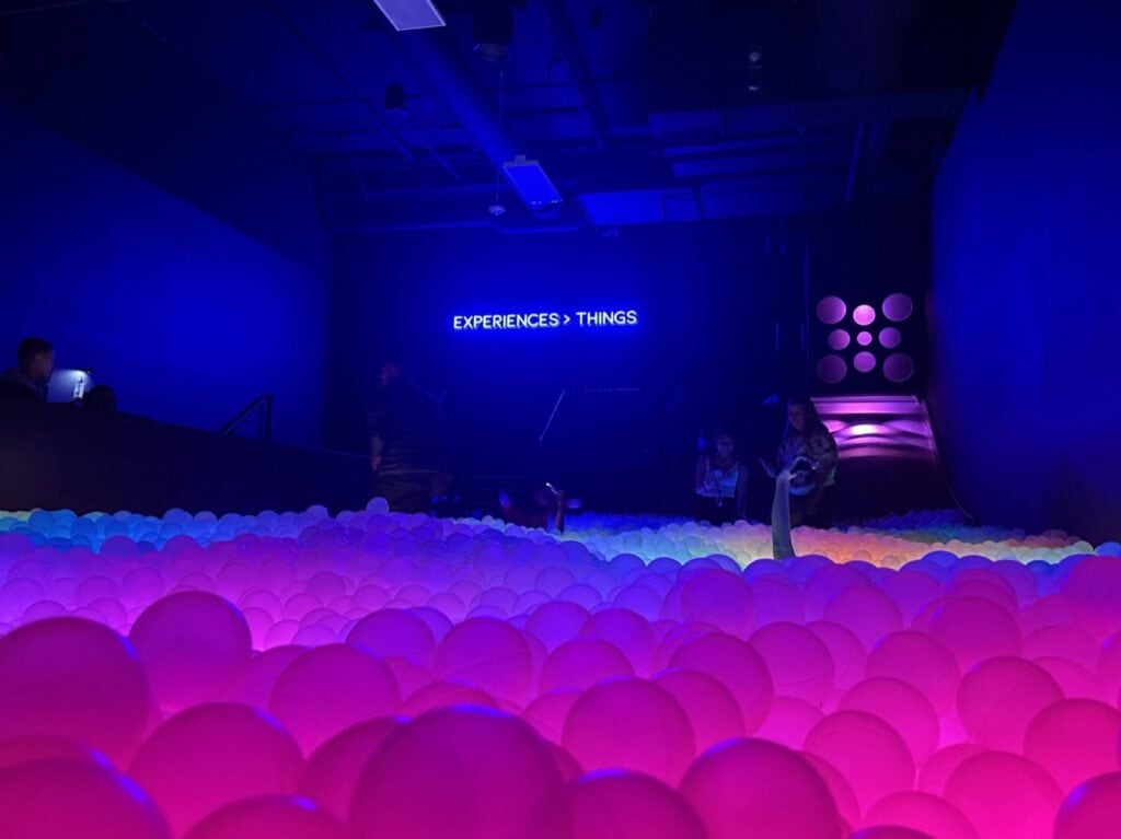 A Dimly Lit Room With People Exploring A Wide Ball Pit Filled With Glowing Pink And Purple Balls. A Neon Sign On The Back Wall Reads Experiences &Gt; Things. The Atmosphere Is Futuristic And Immersive.