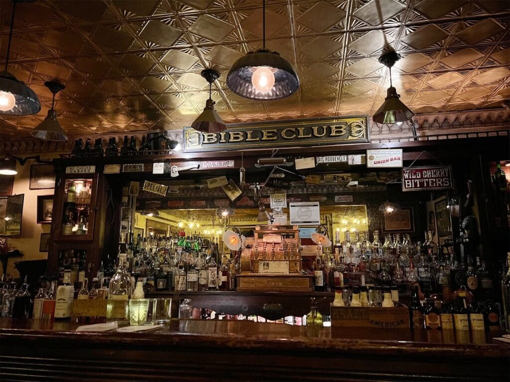 Bible Club'S Cozy Prohibition-Era Bar Interior With A Vintage Vibe Featuring A Counter Lined With Various Bottles Of Spirits. A Sign That Reads &Quot;Bible Club&Quot; Is Prominently Displayed Above The Bar, Surrounded By Warm Lighting, Decorative Hanging Lamps And A Gold Plated Ceiling.