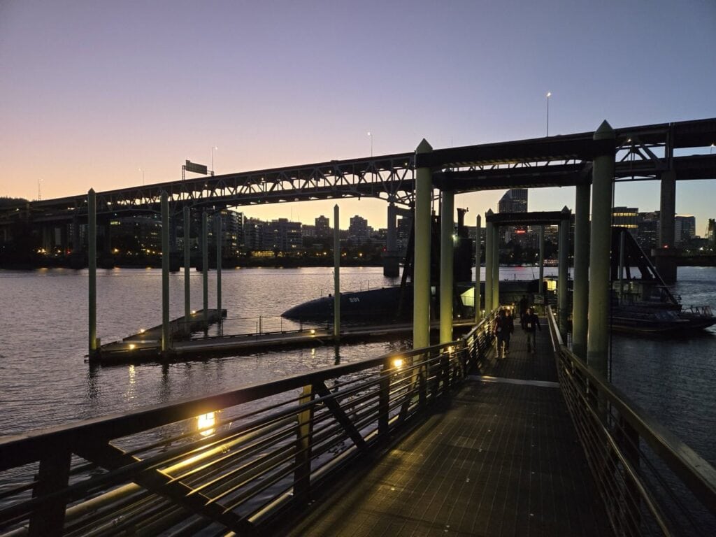 A Dock At Twilight With A View Of A Bridge Spanning A Calm River. Soft Lighting Creates Reflections On The Water, And City Buildings Are Silhouetted Against The Sky. A Few People Are Walking Towards A Docked Submarine.