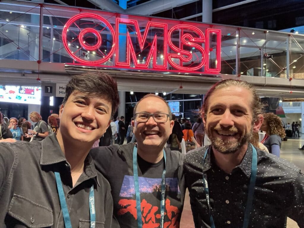 Vinícius, Scott And Max Smile In Front Of A Large Neon Red &Quot;Omsi&Quot; Sign At The Wordcamp Us Afterparty. Other Wordcamp Attendees Are Socializing In The Background.
