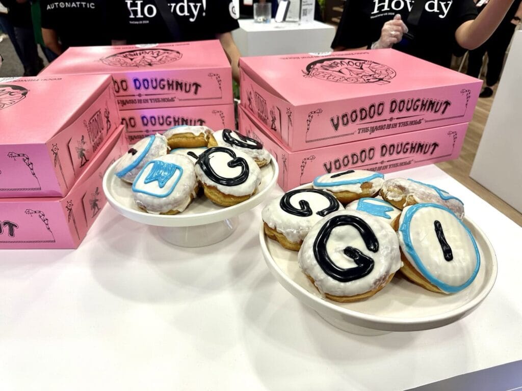 Plates Of Doughnuts With White Icing And Black And Blue Letters Are Displayed On A Table. Behind Them Are Stacks Of Pink Boxes Labeled &Quot;Voodoo Doughnut.&Quot; People In Black T-Shirts Stand In The Background.