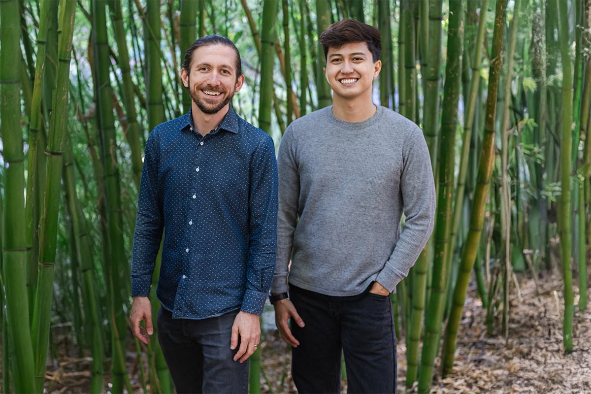 Two people stand smiling in front of tall green bamboo stalks. One person wears a blue patterned shirt and dark jeans, while the other wears a gray sweater and black pants. The ground is covered with dried bamboo leaves.