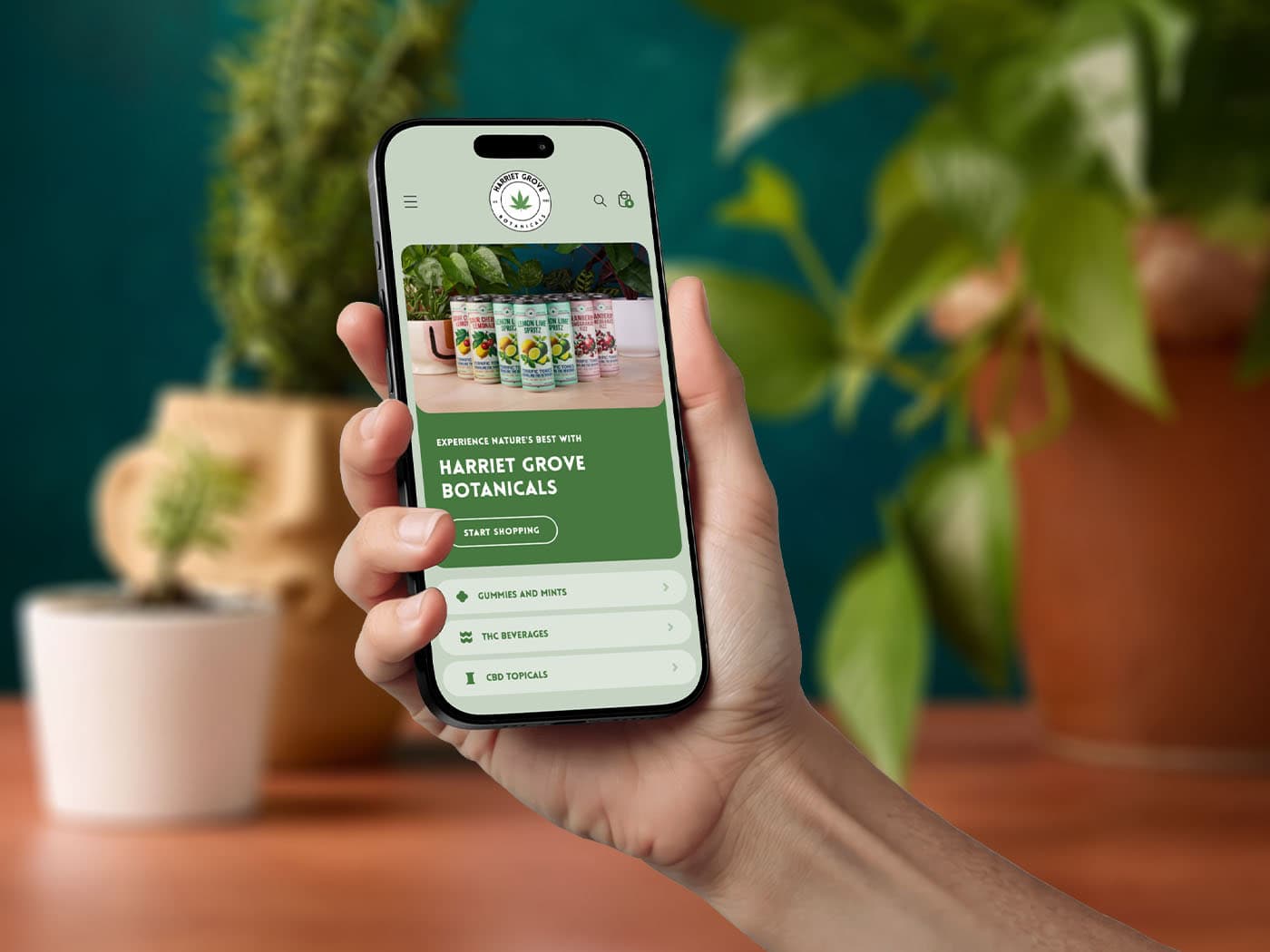A hand holds a smartphone displaying a website for Harriet Grove Botanicals, featuring plant-based products. Blurred potted plants are visible in the background on a wooden surface.