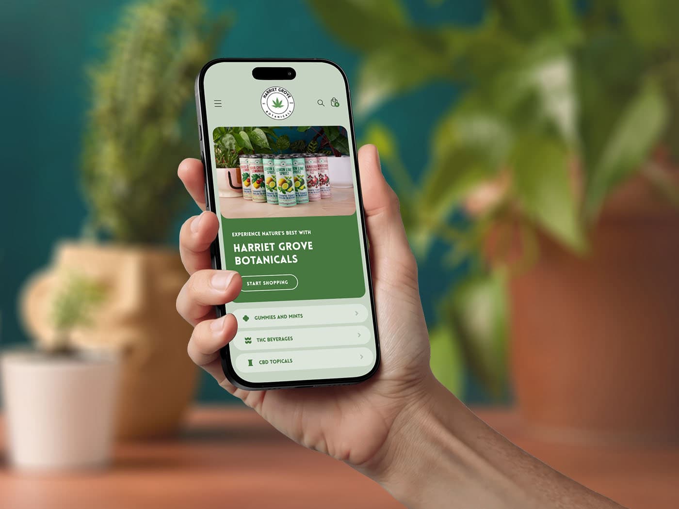 A hand holds a smartphone displaying the Harriet Grove Botanicals website, featuring colorful products and a green, plant-themed design; blurred potted plants are visible in the background.