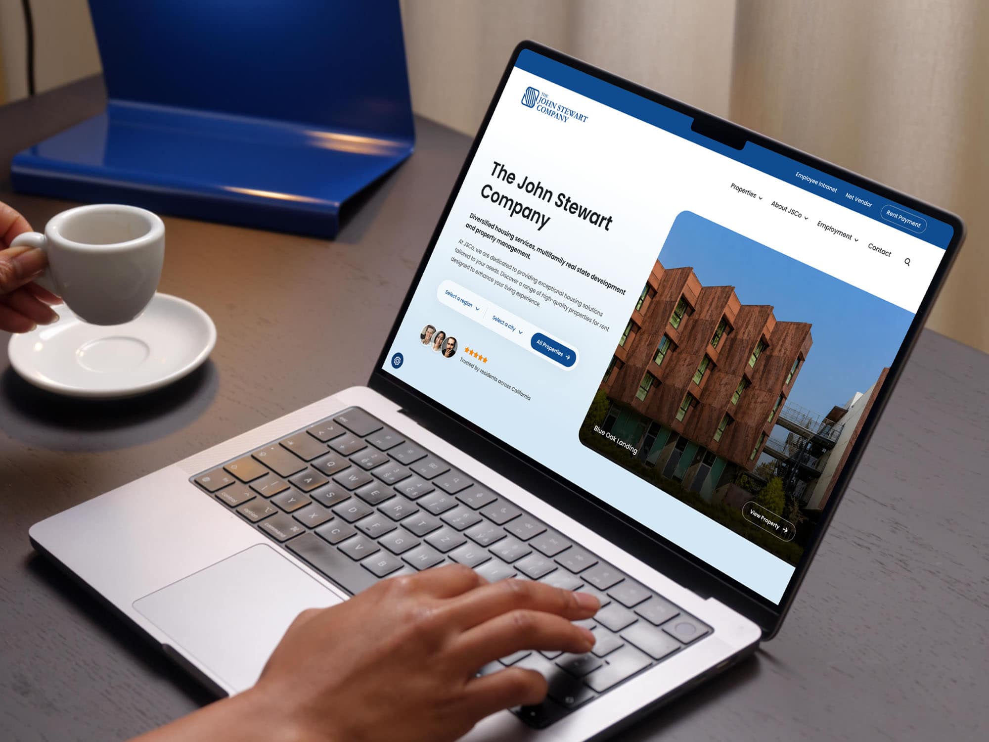 A Person Uses A Laptop Displaying A Website For “The John Stewart Company,” Featuring A Building Photo And Company Information. A Coffee Cup And A Blue Folder Are On The Dark Desk.