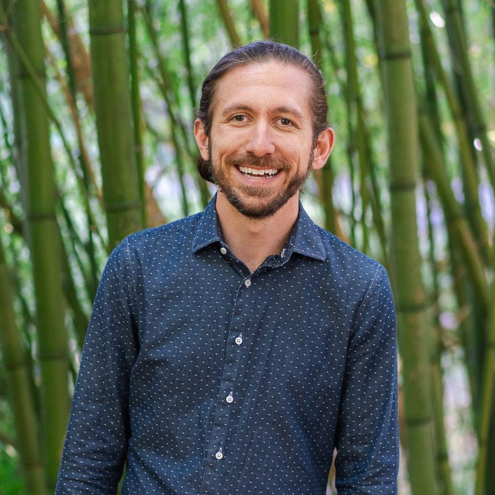 A Man With Long Brown Hair And A Beard, Wearing A Navy Blue, Polka-Dotted Shirt, Smiles While Standing In Front Of Tall Green Bamboo Stalks.