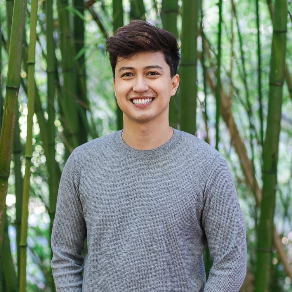 A Smiling Person With Short Brown Hair Wearing A Gray Sweater Stands In Front Of Tall Green Bamboo Stalks In An Outdoor Setting.