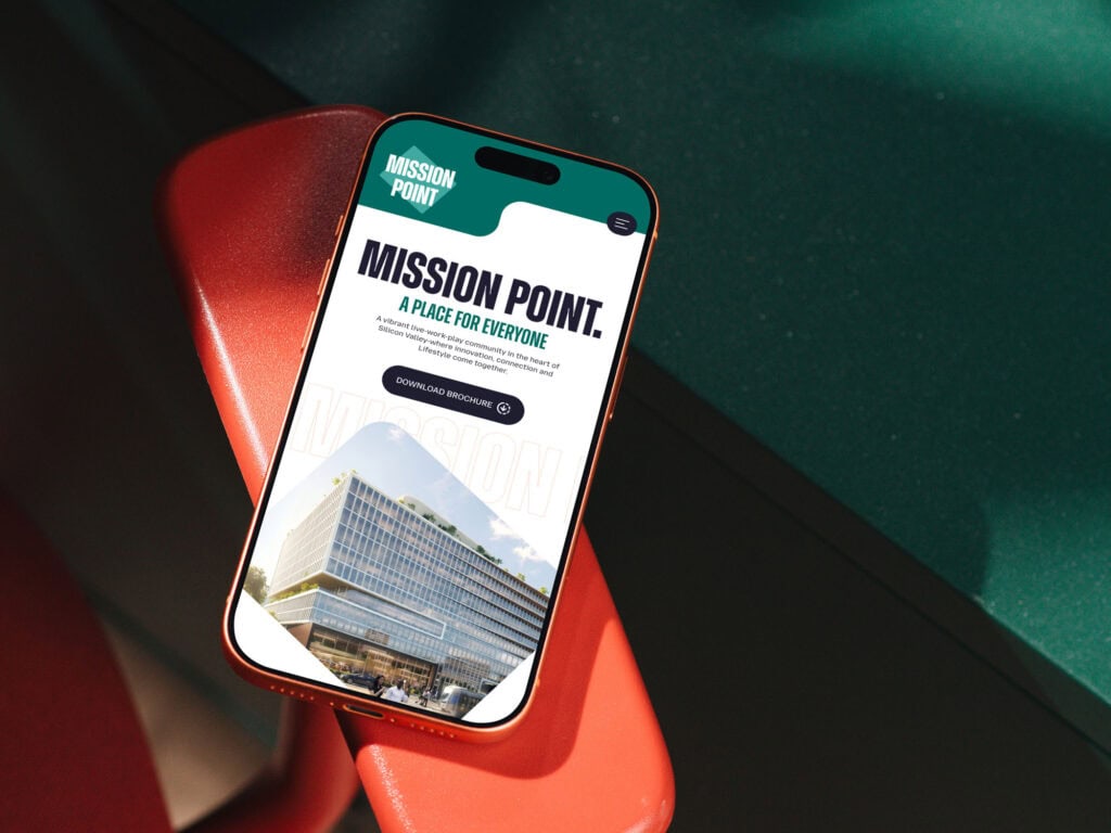 A Smartphone Displaying A Website For Mission Point, Featuring Text, A Building Photo, And A “Download Brochure” Button, Rests On A Red Chair With Green Surfaces In The Background.