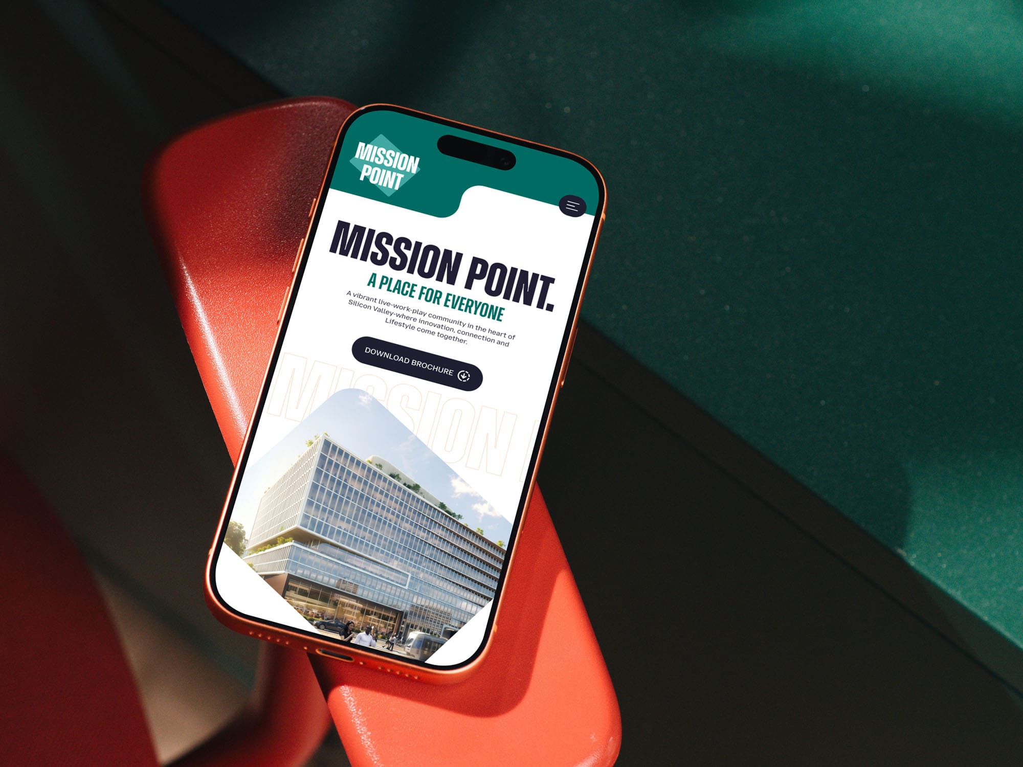 A Smartphone Displaying A Website For Mission Point, Featuring Text, A Building Photo, And A “Download Brochure” Button, Rests On A Red Chair With Green Surfaces In The Background.