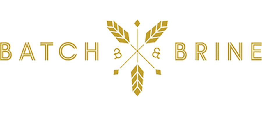 Logo for Batch & Brine featuring golden text with two crossed wheat stalks and arrows in the center, stylized ampersands between the words. White background.