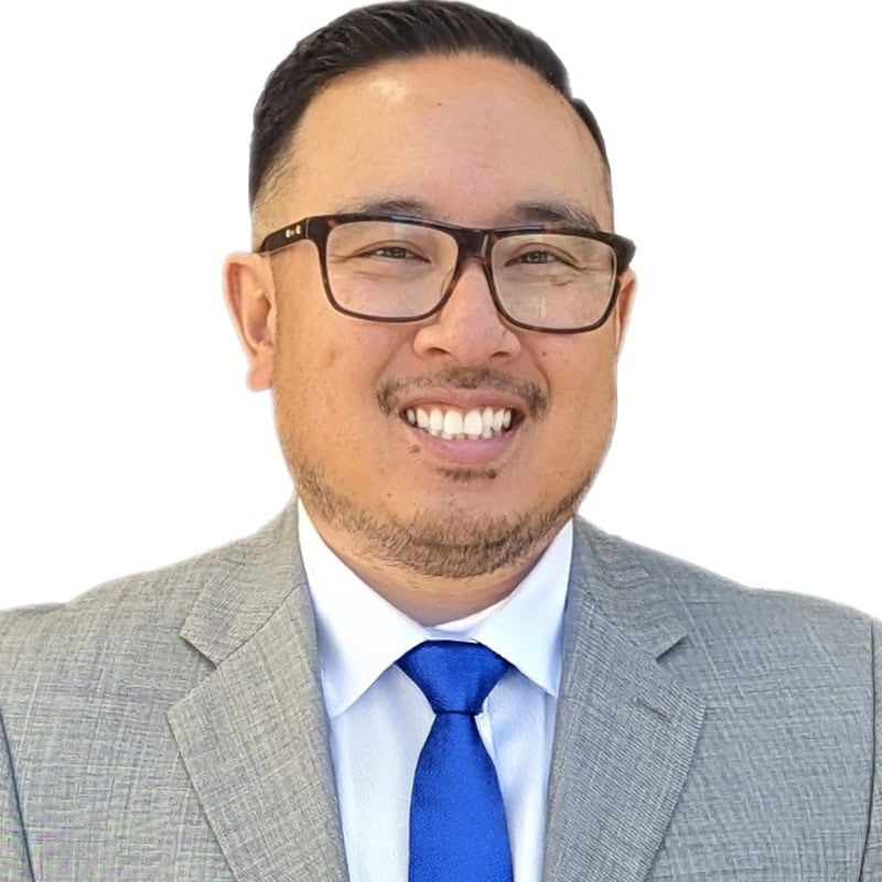Man Wearing Glasses, A Grey Suit, White Shirt, And Blue Tie, Smiling At The Camera Against A Plain White Background.