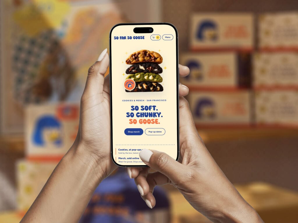 A Person Holds A Smartphone Displaying The So Far So Goose Website, Which Features A Stack Of Four Cookies And Buttons To Shop Merch And View Pop-Up Dates.