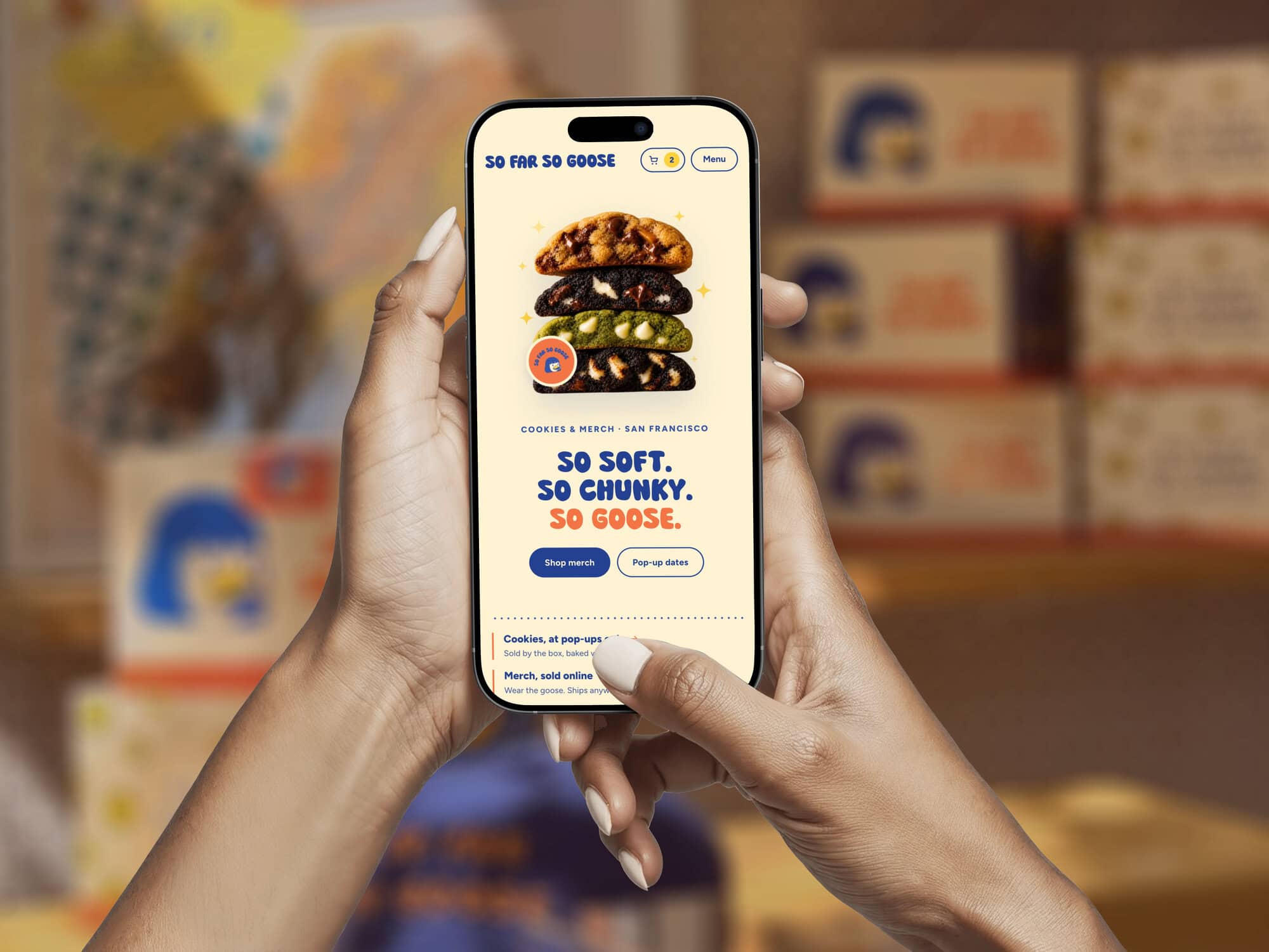 A Person Holds A Smartphone Displaying The So Far So Goose Website, Which Features A Stack Of Four Cookies And Buttons To Shop Merch And View Pop-Up Dates.